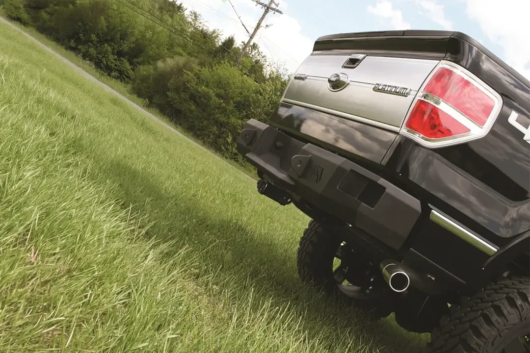 Fab Fours Premium rear bumper on a black Ford F-150, low angled close-up of the rear corner on a grassy slope, showing the heavy-duty bumper end, logo cutouts and hitch area