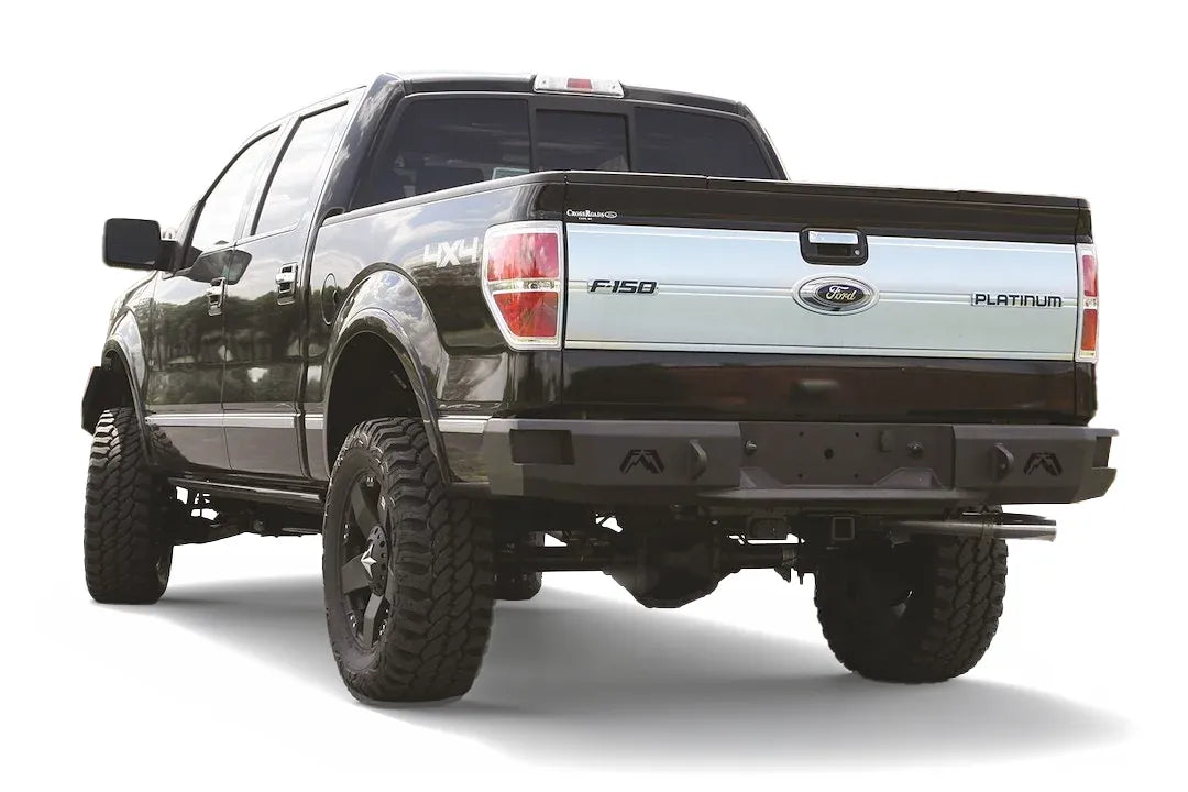 Fab Fours Premium rear bumper on a black Ford F-150, rear driver-side three-quarter view on a white background, showing the heavy-duty bumper, logo cutouts and hitch receiver