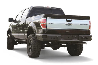 Fab Fours Premium rear bumper on a black Ford F-150, rear driver-side three-quarter view on a white background, showing the heavy-duty bumper, logo cutouts and hitch receiver