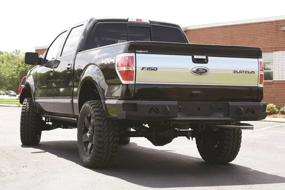 Fab Fours Premium rear bumper on a black Ford F-150, rear driver-side three-quarter view in a parking lot, showing the heavy-duty bumper, logo cutouts and hitch receiver