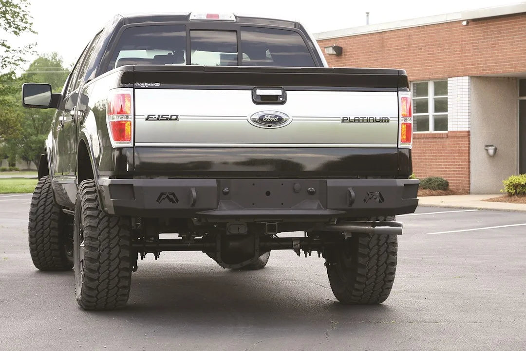 Fab Fours Premium rear bumper on a black Ford F-150, rear view in a parking lot, showing the heavy-duty bumper, logo cutouts and center plate area
