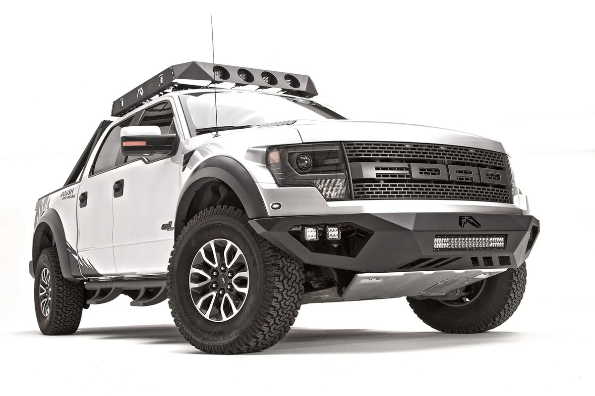 Fab Fours Vengeance front bumper on a silver Ford Raptor, front passenger-side three-quarter studio view, showing the sculpted bumper, light bar and cube light pods