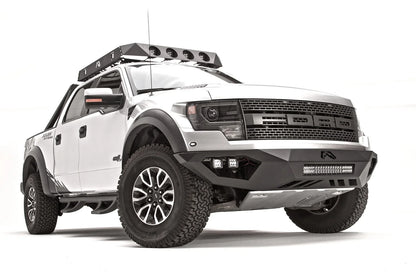 Fab Fours Vengeance front bumper on a silver Ford Raptor, front passenger-side three-quarter studio view, showing the sculpted bumper, light bar and cube light pods