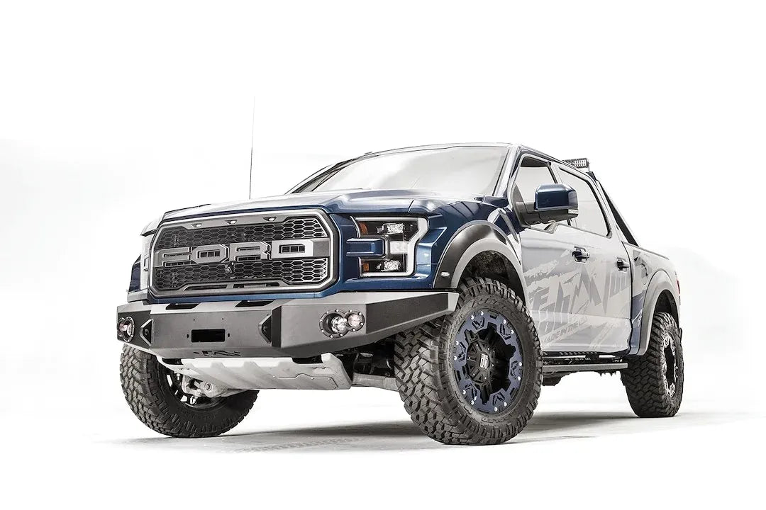Fab Fours Premium Winch front bumper on a blue and gray wrapped Ford Raptor, front driver-side three-quarter view, showing the winch recess, round fog lights and shackle mounts