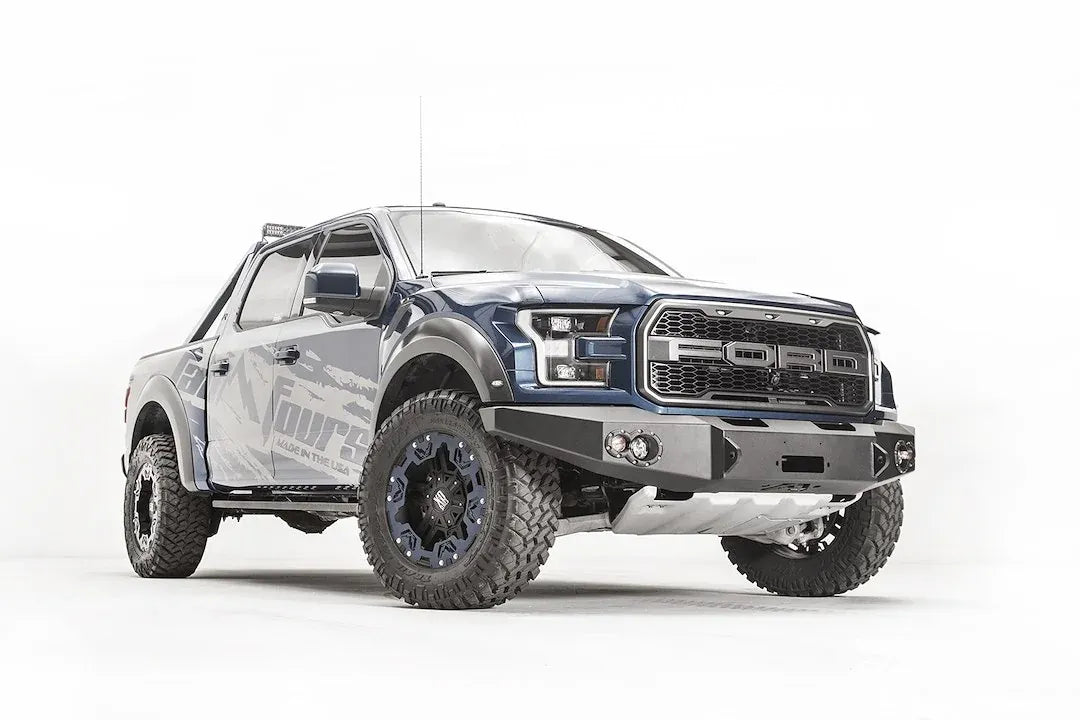 Fab Fours Premium Winch front bumper on a blue and gray wrapped Ford Raptor, front passenger-side three-quarter view, showing the winch recess, round fog lights and bumper ends