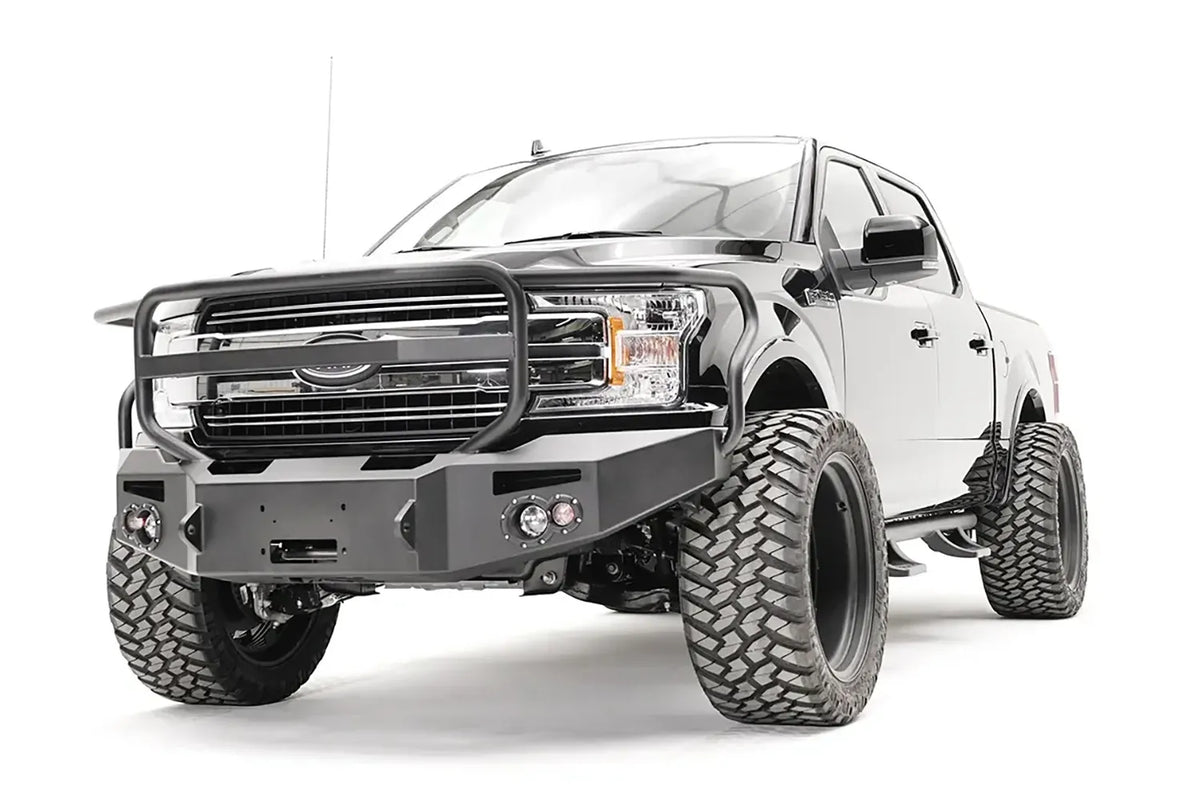 Fab Fours Premium Winch front bumper with full guard on a black Ford F-150, front driver-side three-quarter view, showing the full grille guard, winch recess and round fog lights