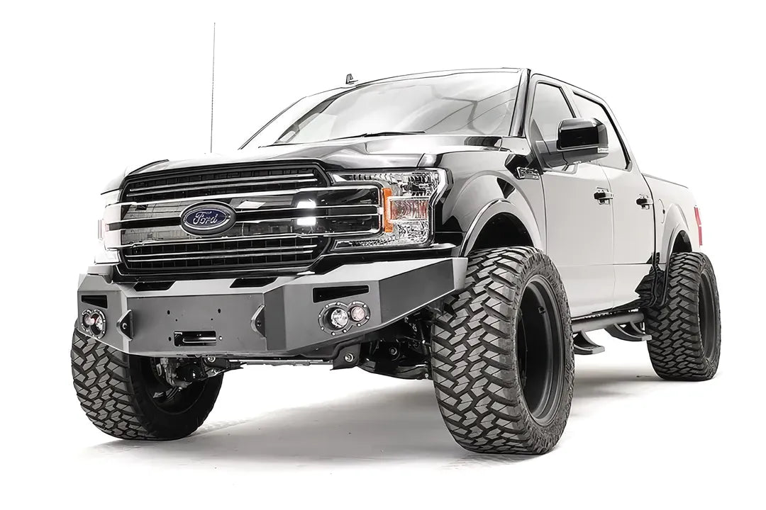 Fab Fours Premium Winch front bumper on a black Ford F-150, front driver-side three-quarter view, showing the winch recess, round fog lights and bumper ends