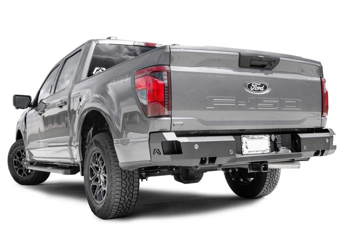 Fab Fours Premium rear bumper on a gray Ford F-150, rear driver side view, showing the sensor cutouts, the corner step pads and the integrated hitch receiver opening