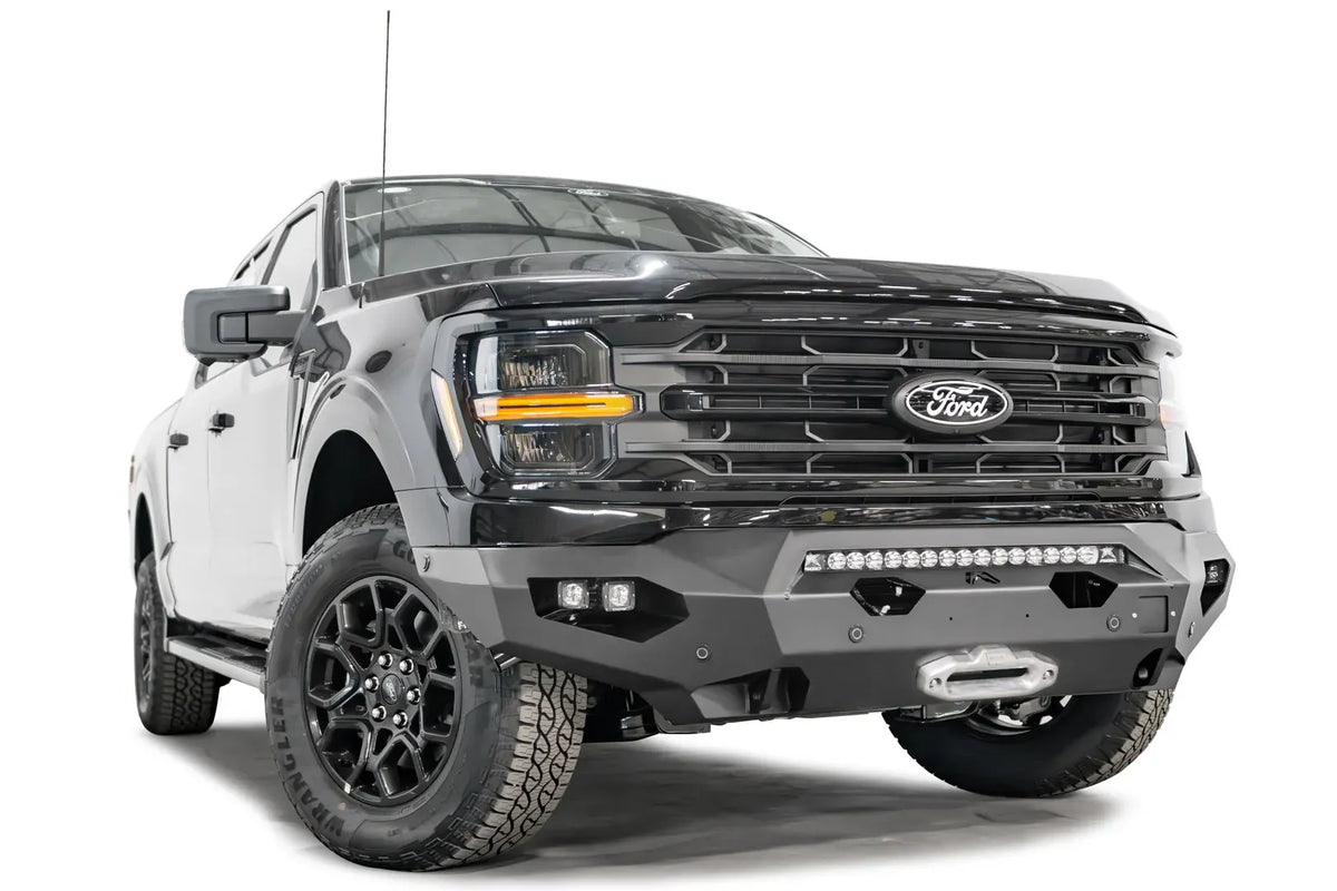 Fab Fours Matrix front bumper on a black Ford F-150, front driver-side three-quarter view, showing the light bar, winch fairlead and cube light pods