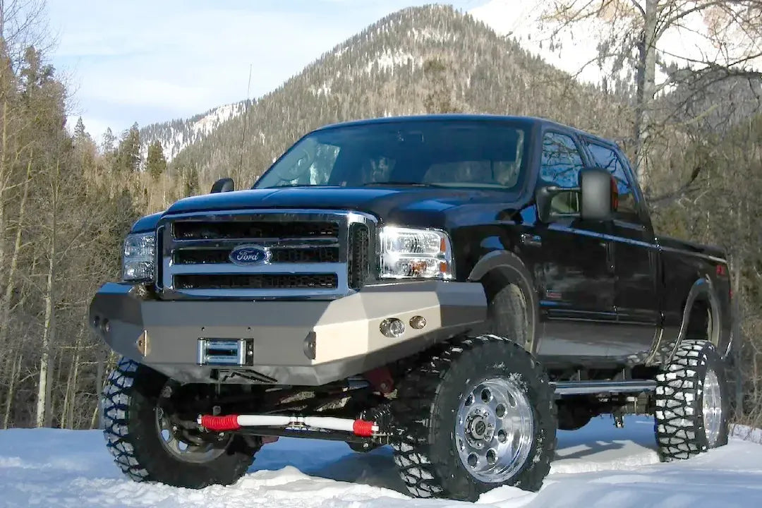 Fab Fours Premium Heavy Duty winch front bumper on a black Ford Super Duty in a snowy mountain setting, front driver-side three-quarter view, showing the winch opening, integrated lights and shackle mounts