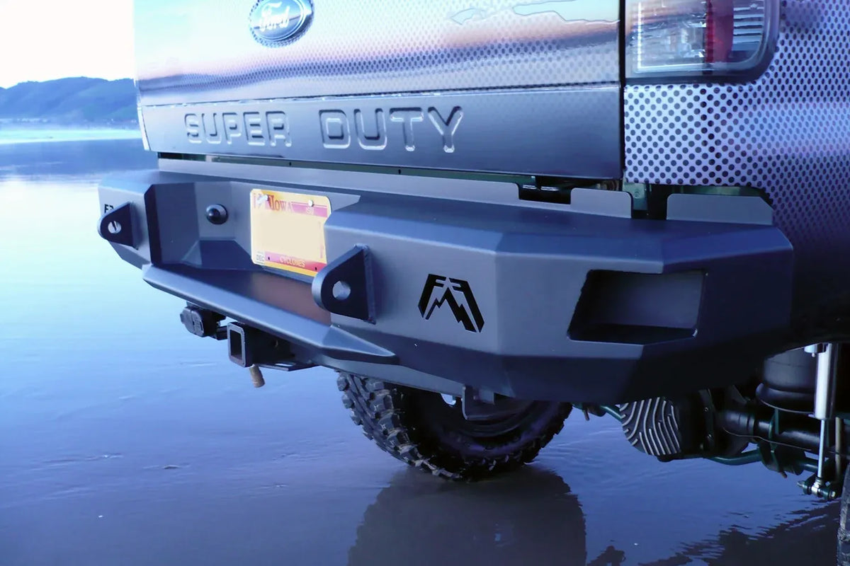Fab Fours Premium rear bumper on a dark Ford Super Duty outdoors, close-up of the rear corner, showing the Fab Fours logo cutout, shackle mount and hitch receiver