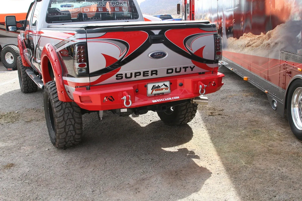 Fab Fours Premium rear bumper in red on a wrapped Ford Super Duty outdoors, rear driver-side three-quarter view, showing the bumper face, D-ring shackles and hitch receiver