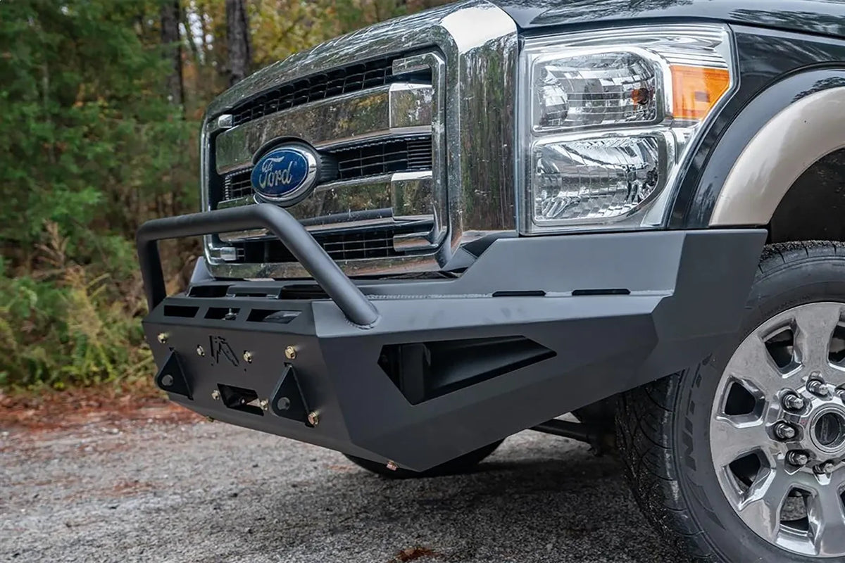 Fab Fours Red Steel front bumper with pre-runner guard on a dark green and tan Ford Super Duty outdoors, close-up of the front corner, showing the pre-runner tube, shackle tabs and angular wing cutout