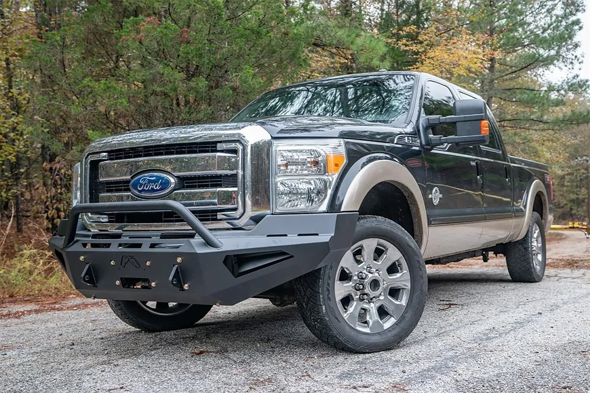 Fab Fours Red Steel front bumper with pre-runner guard on a dark green and tan Ford Super Duty outdoors, front driver-side three-quarter view, showing the pre-runner hoop, winch plate and angular wing cutouts