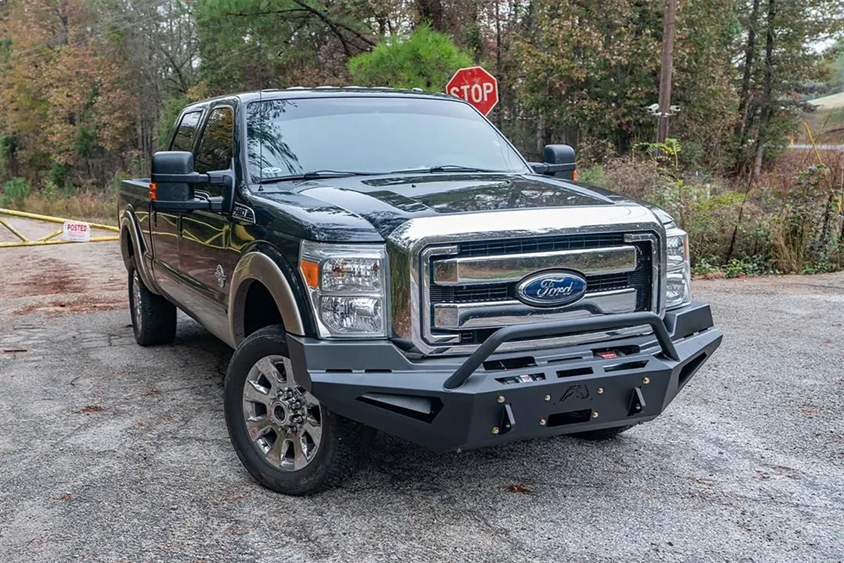 Fab Fours Red Steel front bumper with pre-runner guard on a dark green and tan Ford Super Duty outdoors, front passenger-side three-quarter view, showing the tubular pre-runner hoop, winch plate and angular wings