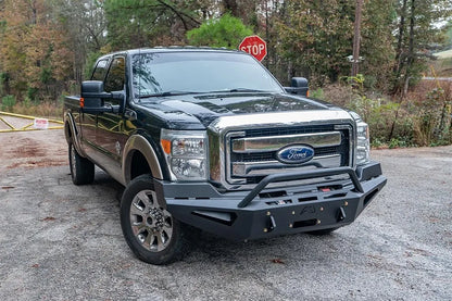 Fab Fours Red Steel front bumper with pre-runner guard on a dark green and tan Ford Super Duty outdoors, front passenger-side three-quarter view, showing the tubular pre-runner hoop, winch plate and angular wings