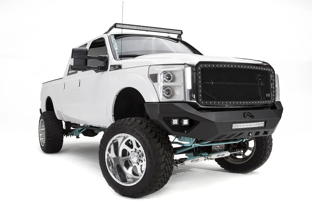 Fab Fours Vengeance front bumper on a white Ford Super Duty, front passenger-side three-quarter view, showing the center light bar, pod lights and angular bumper wing