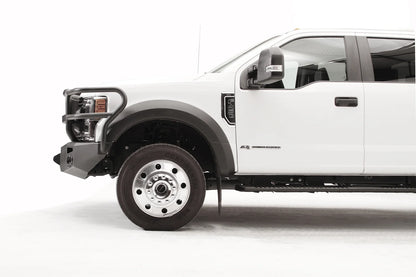 Fab Fours Premium Winch front bumper with full guard on a white Ford Super Duty F-450/F-550, driver-side profile view of the front corner, showing full grille guard hoops, the center winch opening, round light cutouts, and angular steel end caps