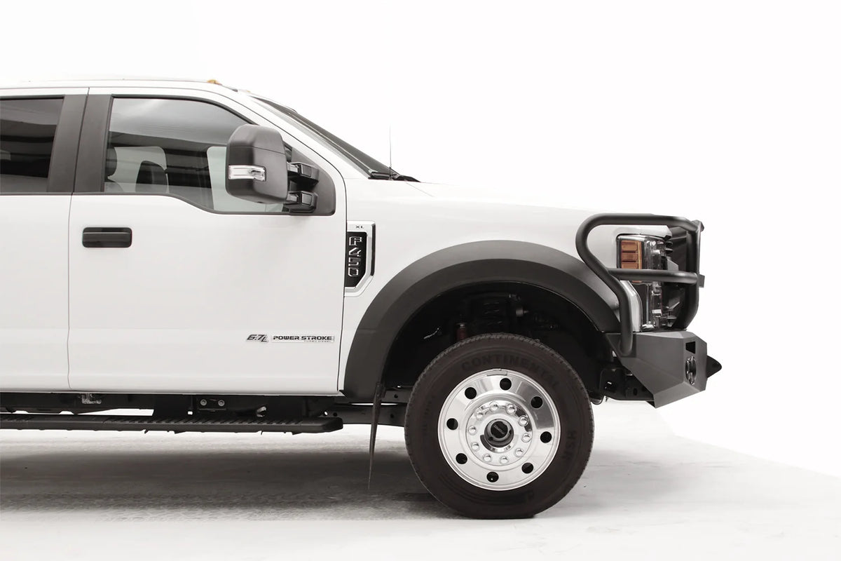 Fab Fours Premium Winch front bumper with full guard on a white Ford Super Duty F-450/F-550, passenger-side profile view of the front corner, showing full grille guard hoops, the center winch opening, round light cutouts, and angular steel end caps