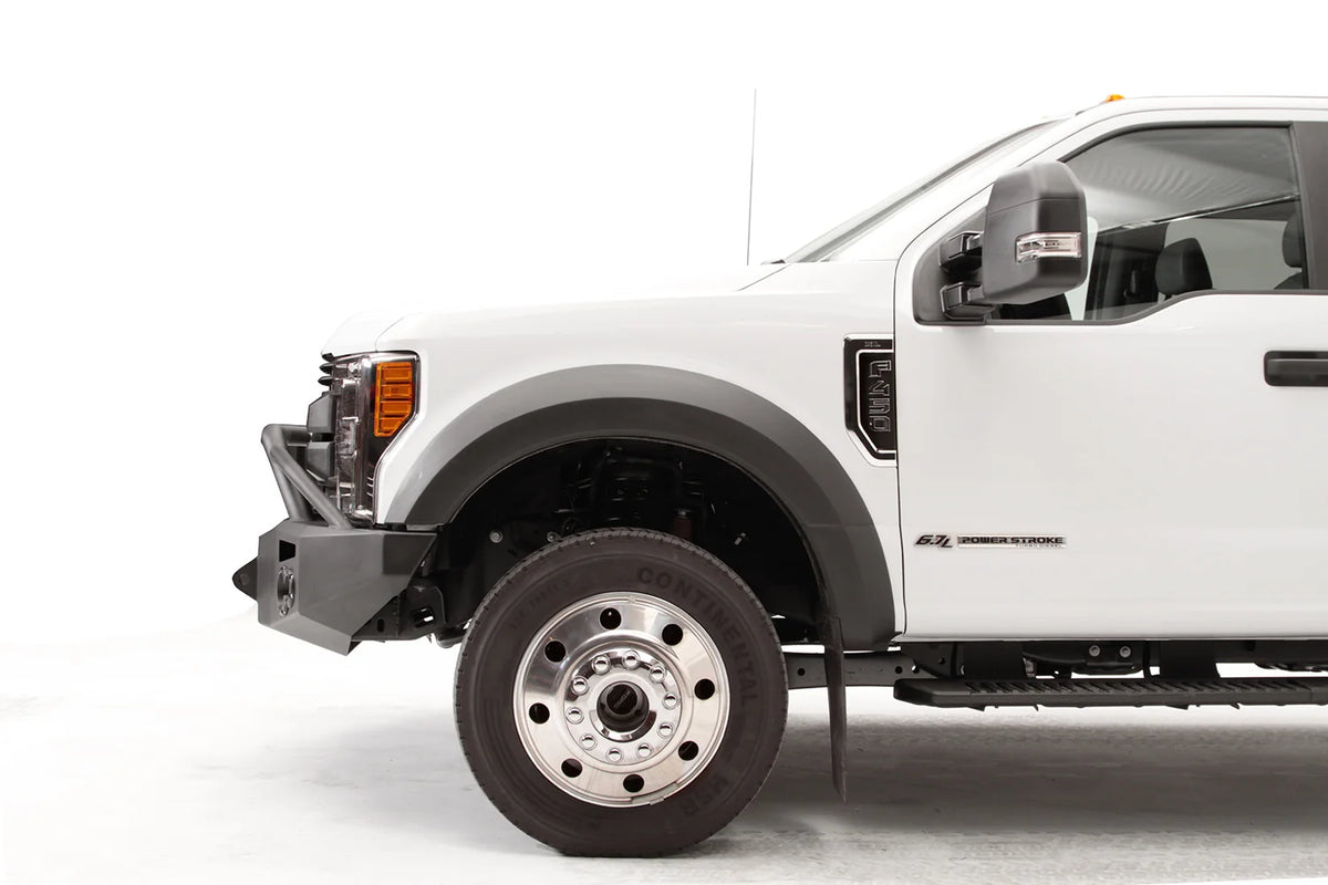 Fab Fours Premium Winch front bumper with pre-runner guard on a white Ford Super Duty F-450/F-550, driver-side profile view of the front corner, showing the tubular pre-runner hoop, the center winch opening, round light cutouts, and angular steel end caps