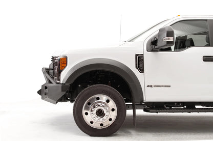 Fab Fours Premium Winch front bumper with pre-runner guard on a white Ford Super Duty F-450/F-550, driver-side profile view of the front corner, showing the tubular pre-runner hoop, the center winch opening, round light cutouts, and angular steel end caps