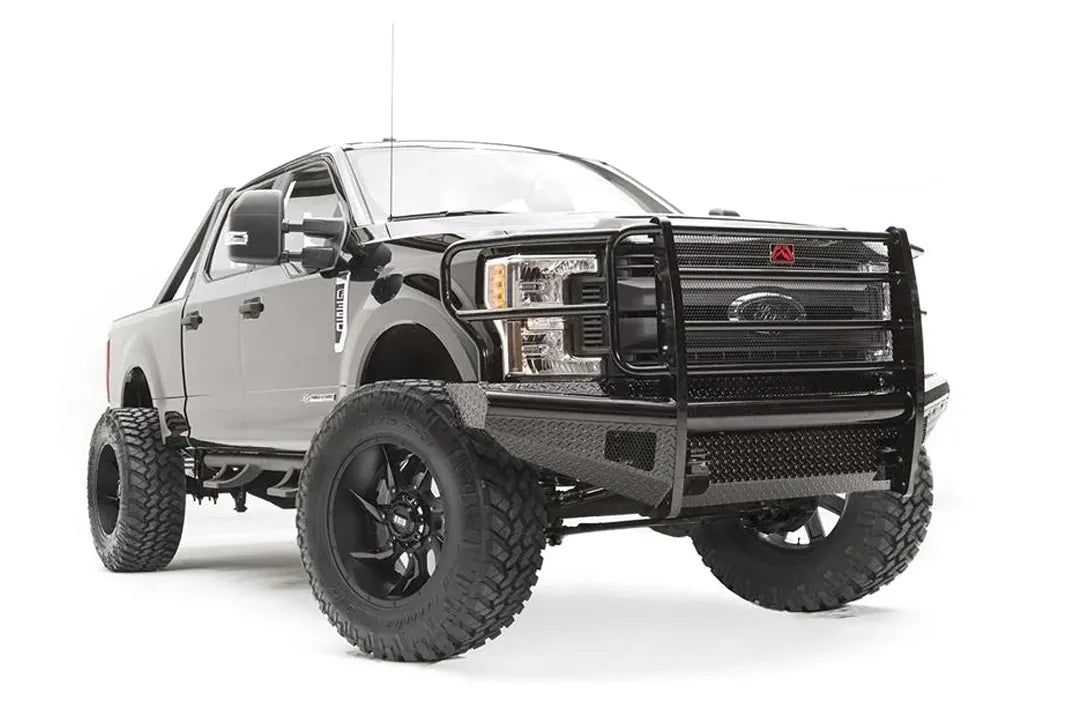 Fab Fours Black Steel front bumper with full guard on a black Ford Super Duty, front passenger-side three-quarter view, showing full grille guard with mesh insert, diamond-plate face panels, the center license plate mount, and angled side wings