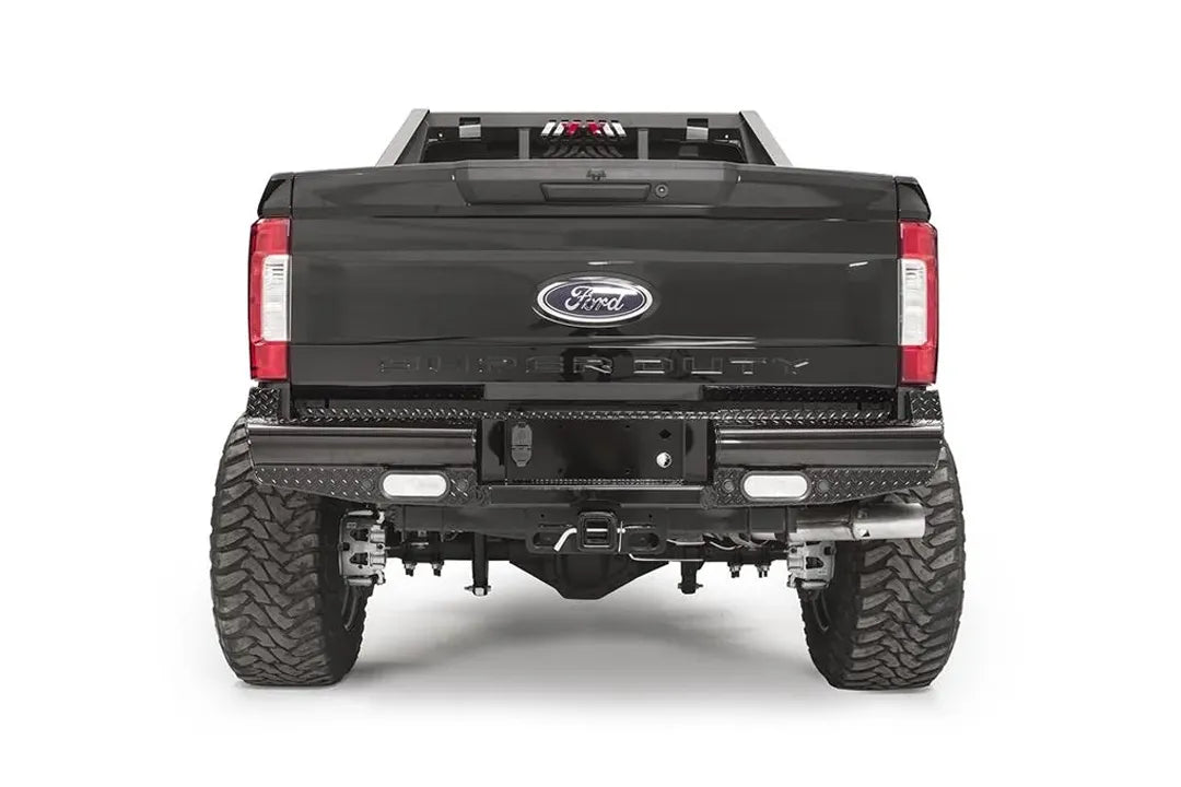 Fab Fours Black Steel rear bumper on a black Ford Super Duty, straight-on rear view, showing diamond-plate top surfaces, integrated backup light openings, and the center receiver hitch access