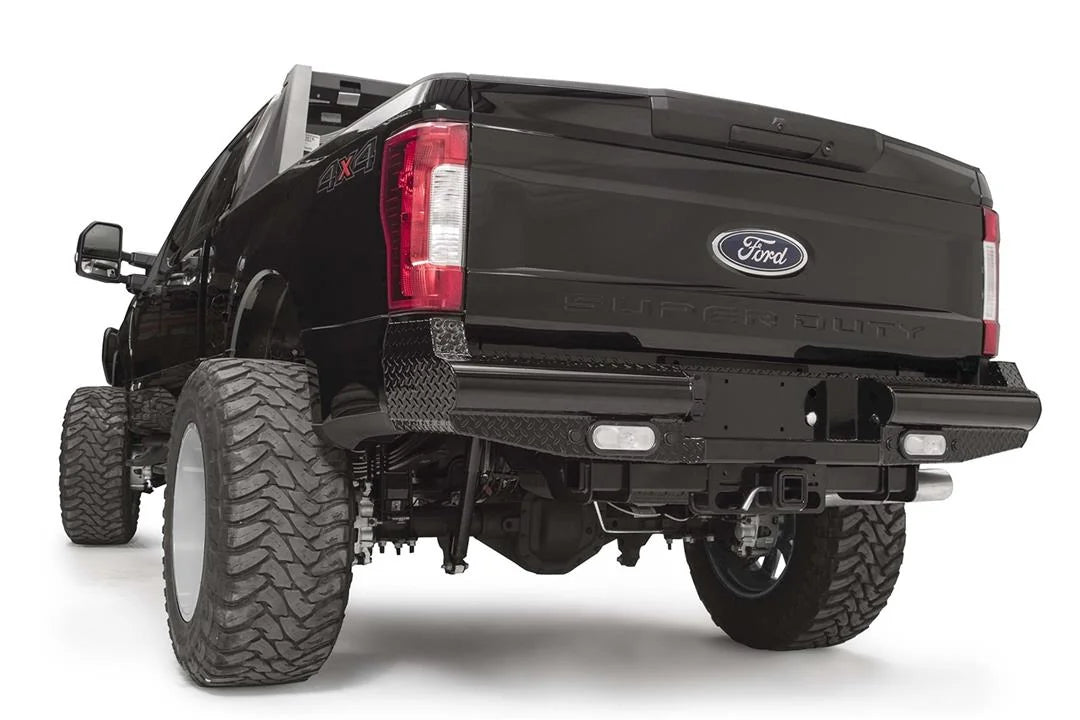 Fab Fours Black Steel rear bumper on a black Ford Super Duty, rear driver-side three-quarter view, showing diamond-plate top surfaces, integrated backup light openings, and the center receiver hitch access