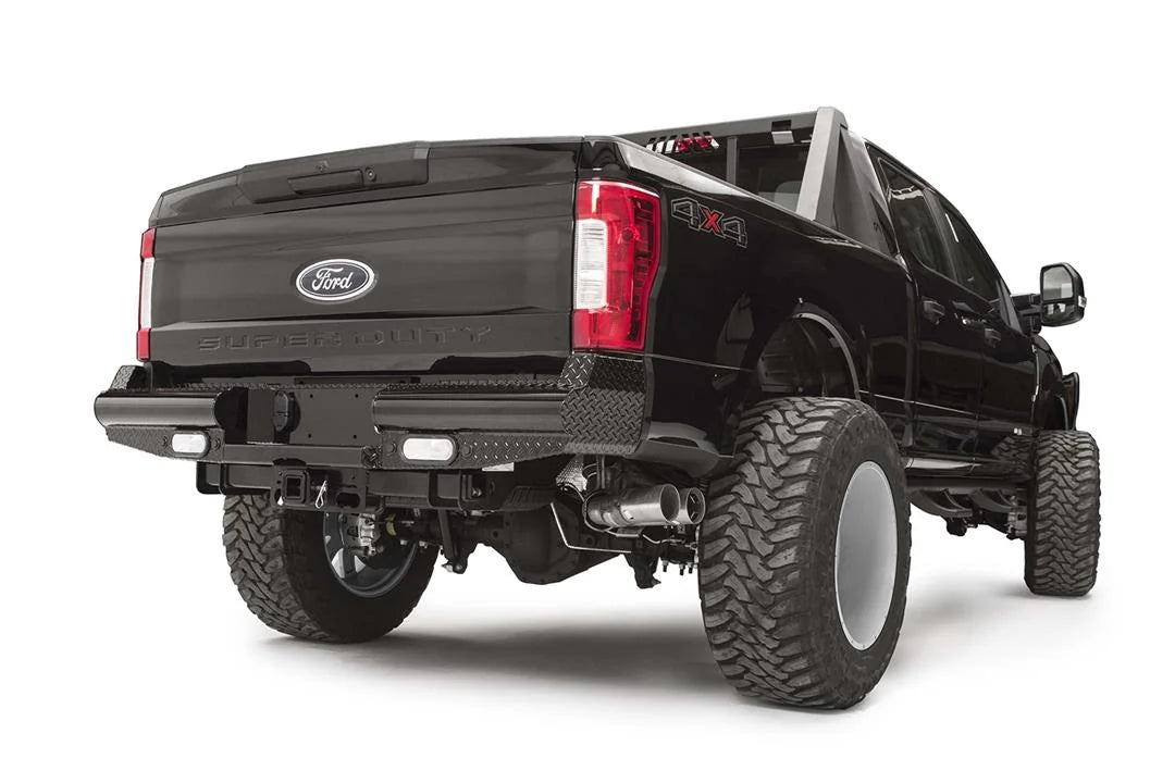 Fab Fours Black Steel rear bumper on a black Ford Super Duty, rear passenger-side three-quarter view, showing diamond-plate top surfaces, integrated backup light openings, and the center receiver hitch access