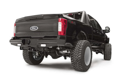 Fab Fours Black Steel rear bumper on a black Ford Super Duty, rear passenger-side three-quarter view, showing diamond-plate top surfaces, integrated backup light openings, and the center receiver hitch access