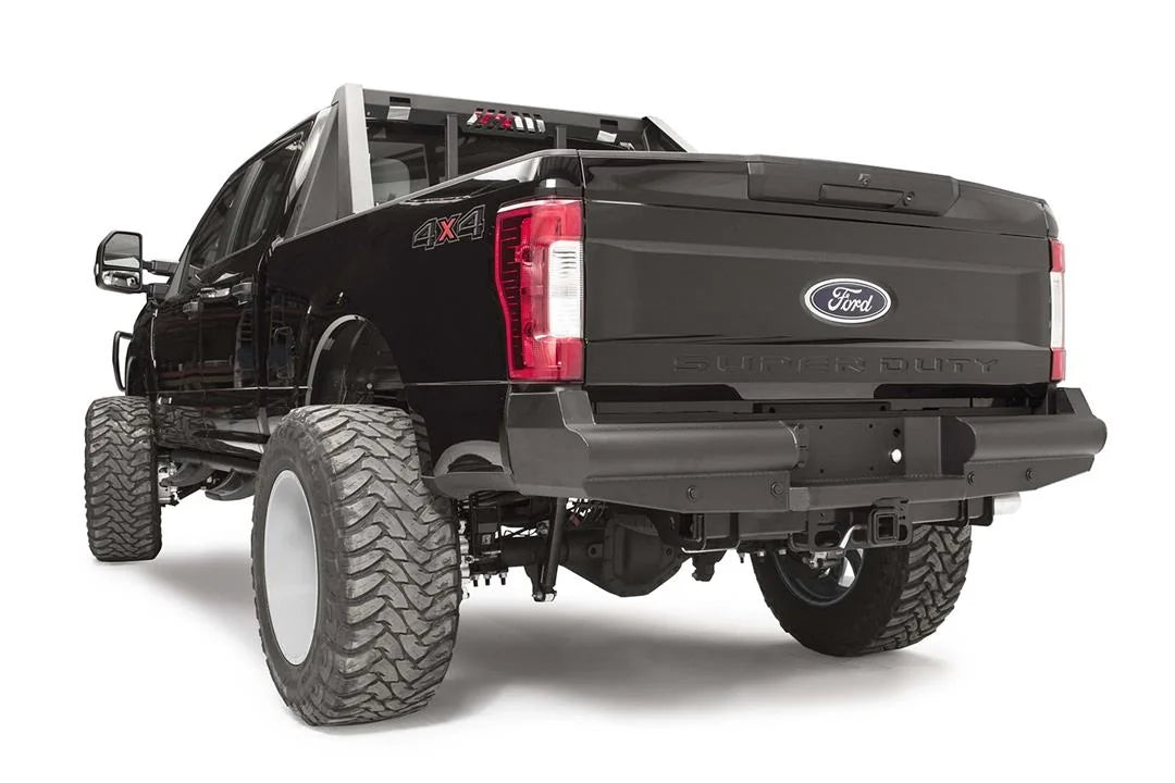 Fab Fours Black Steel Elite rear bumper on a black Ford Super Duty, rear driver-side three-quarter view, showing the smooth steel face, the center license plate recess, and the receiver hitch access