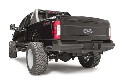 Fab Fours Black Steel Elite rear bumper on a black Ford Super Duty, rear driver-side three-quarter view, showing the smooth steel face, the center license plate recess, and the receiver hitch access