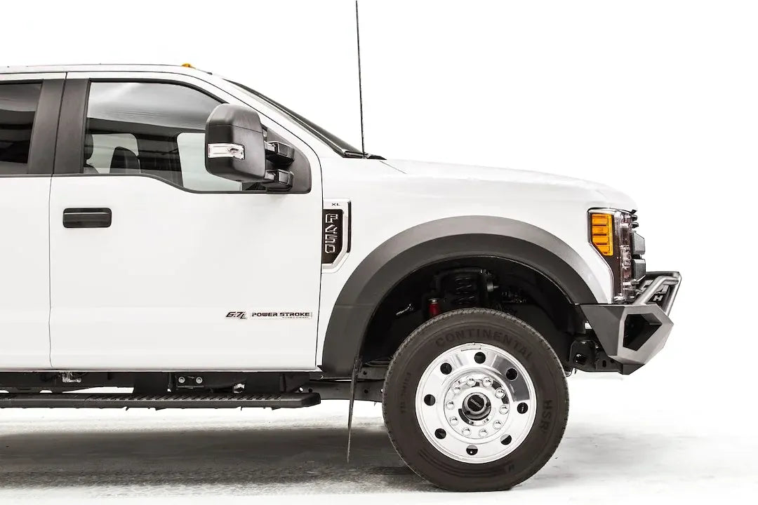 Fab Fours Vengeance front bumper with pre-runner guard on a white Ford Super Duty F-450/F-550, passenger-side profile view of the front corner, showing the tubular pre-runner hoop, the center LED light bar, cube light pods, and the open skid plate design