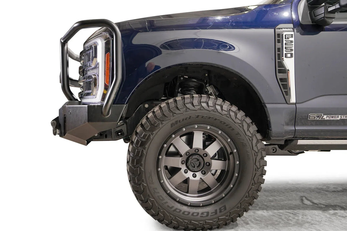 Fab Fours Premium Winch front bumper with full guard on a dark blue Ford Super Duty F-250/F-350, driver-side profile view of the front corner, showing full grille guard hoops, the center winch plate with fairlead, LED light pods, and D-ring shackle mounts
