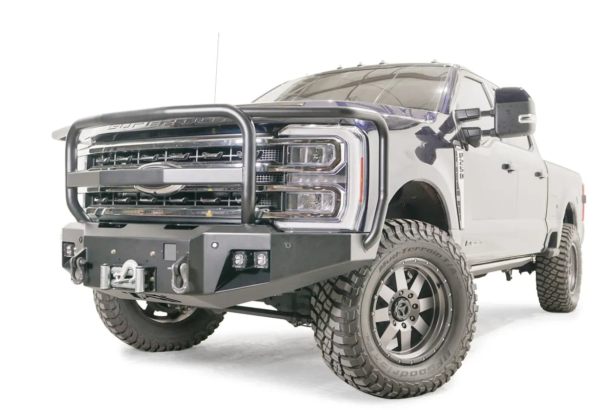 Fab Fours Premium Winch front bumper with full guard on a blue and gray two-tone Ford Super Duty F-250/F-350, front driver-side three-quarter view, showing full grille guard hoops, the center winch plate with fairlead, LED light pods, and D-ring shackle mounts