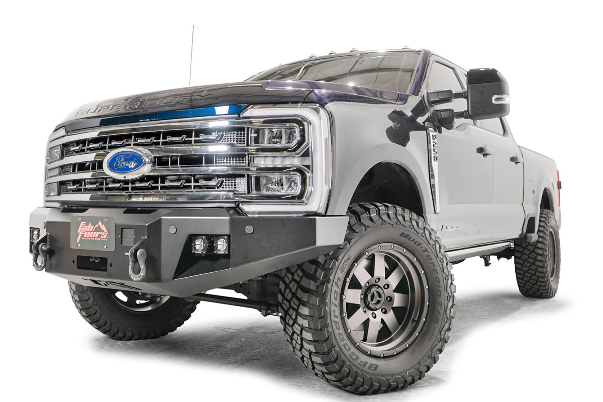 Fab Fours Premium Winch front bumper on a blue and gray two-tone Ford Super Duty F-250/F-350, front driver-side three-quarter view, showing the center winch plate with fairlead, LED light pods, and D-ring shackle mounts