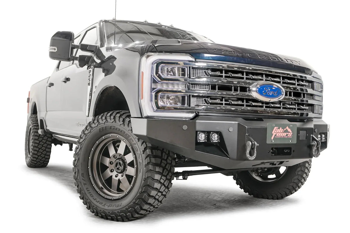 Fab Fours Premium Winch front bumper on a blue and gray two-tone Ford Super Duty F-250/F-350, front passenger-side three-quarter view, showing the center winch plate with fairlead, LED light pods, and D-ring shackle mounts