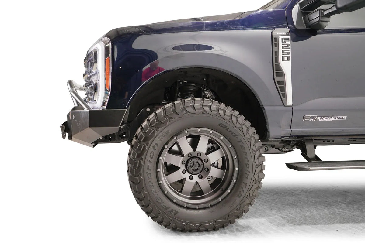 Fab Fours Premium Winch front bumper with pre-runner guard on a dark blue Ford Super Duty F-250/F-350, driver-side profile view of the front corner, showing the tubular pre-runner hoop, the center winch plate with fairlead, LED light pods, and D-ring shackle mounts