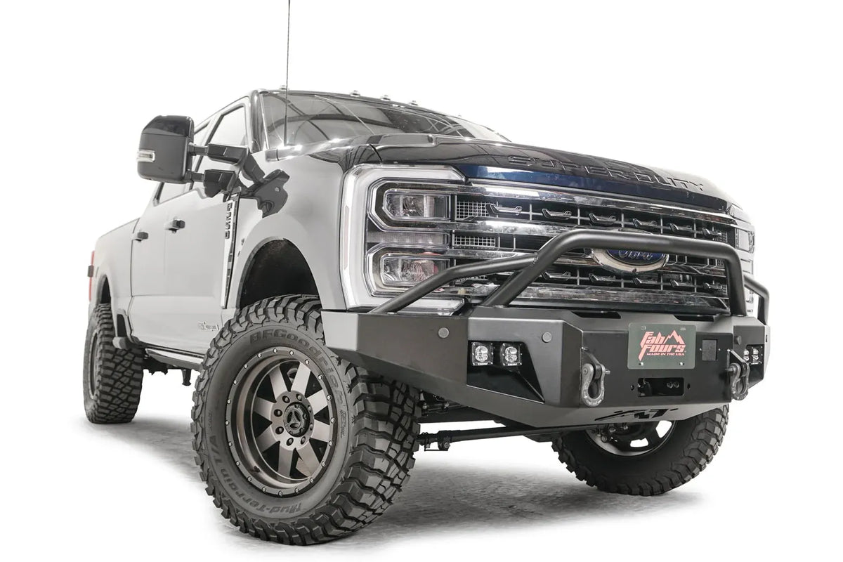 Fab Fours Premium Winch front bumper with pre-runner guard on a blue and gray two-tone Ford Super Duty F-250/F-350, front passenger-side three-quarter view, showing the tubular pre-runner hoop, the center winch plate with fairlead, LED light pods, and D-ring shackle mounts
