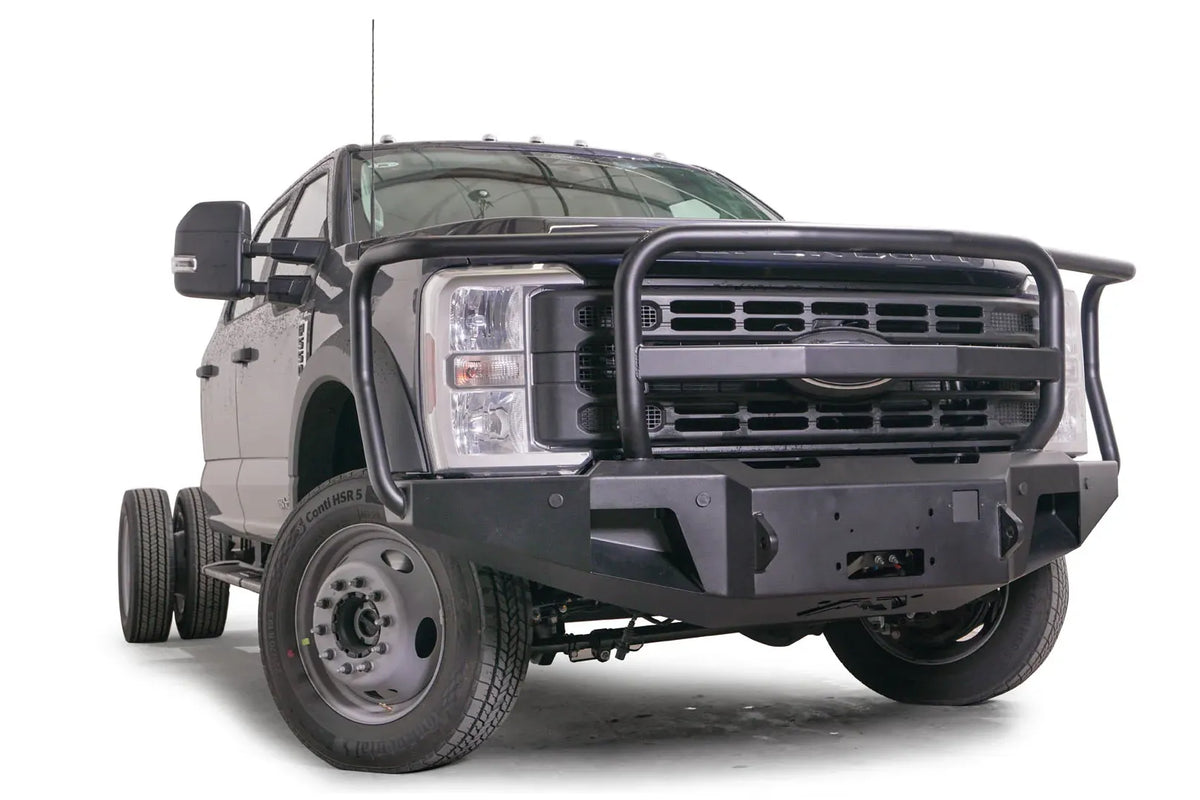 Fab Fours Premium Winch front bumper with full guard on a blue and gray two-tone Ford Super Duty F-450/F-550, front passenger-side three-quarter view, showing full grille guard hoops, the center winch plate, angular end caps, and recessed light openings