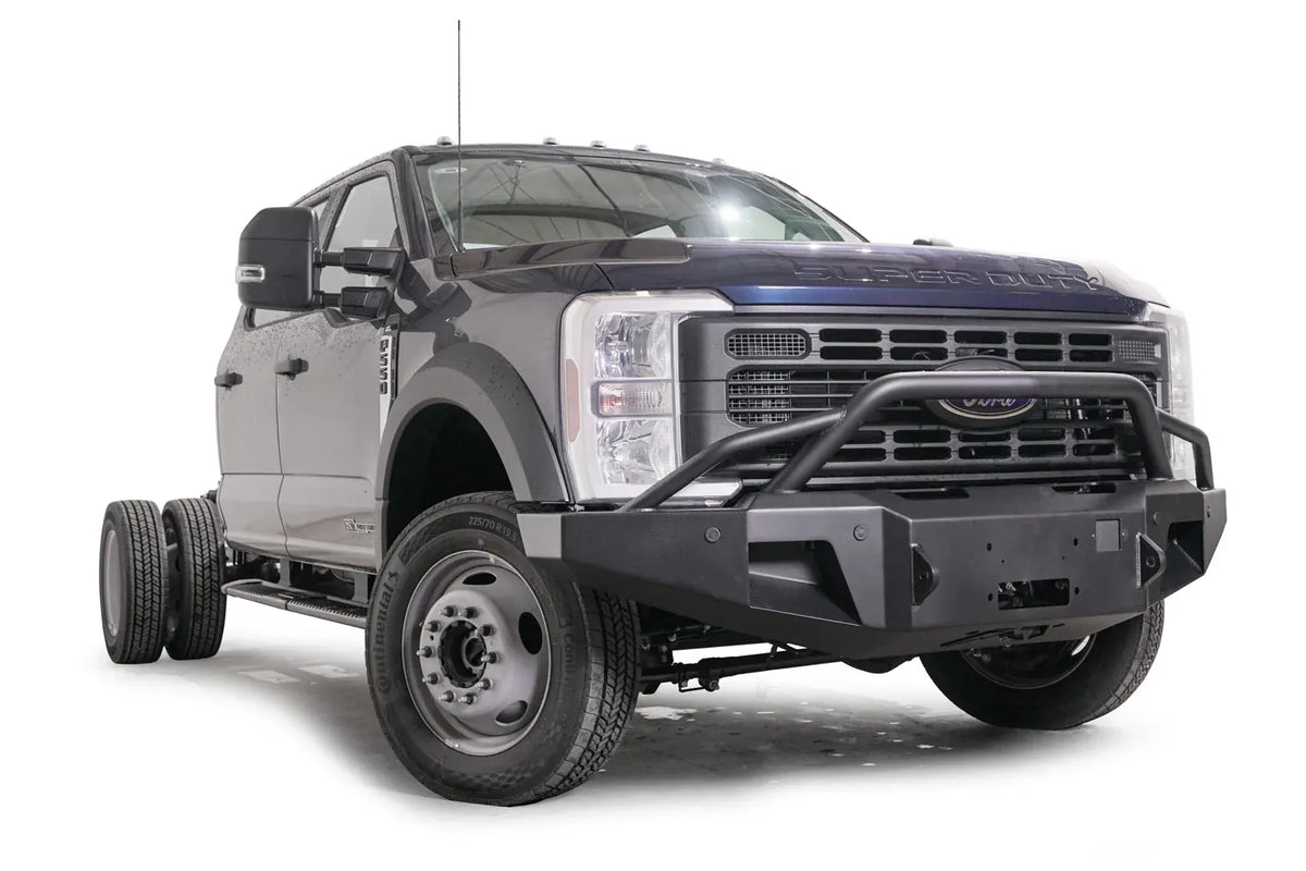 Fab Fours Premium Winch front bumper with pre-runner guard on a blue and gray two-tone Ford Super Duty F-450/F-550, front passenger-side three-quarter view, showing the tubular pre-runner hoop, the center winch plate, angular end caps, and recessed light openings