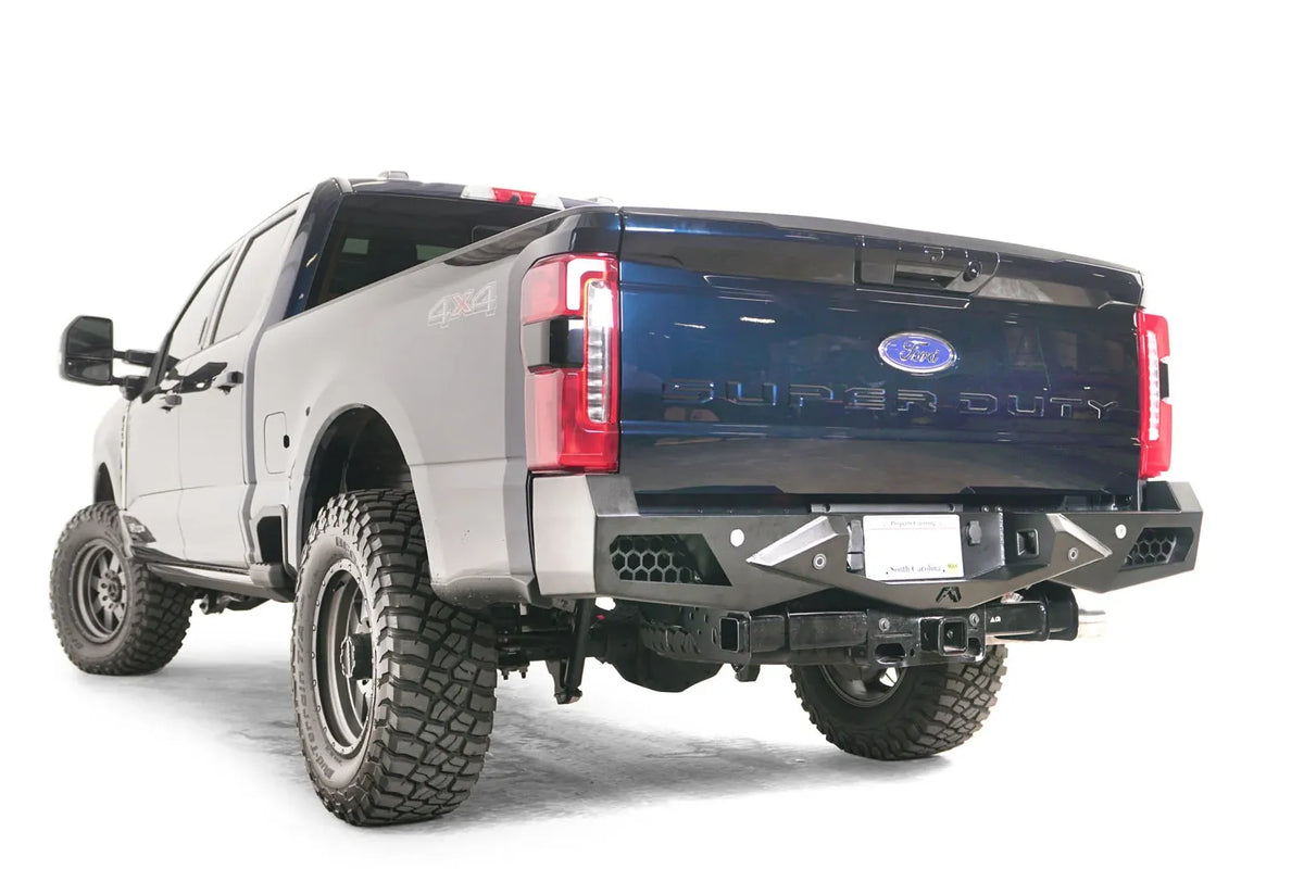 Fab Fours Vengeance rear bumper on a blue and gray two-tone Ford Super Duty, rear driver-side three-quarter view, showing honeycomb-pattern side cutouts, the center license plate recess, and the receiver hitch access