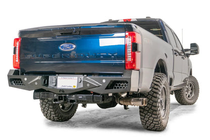 Fab Fours Vengeance rear bumper on a blue and gray two-tone Ford Super Duty, rear passenger-side three-quarter view, showing honeycomb-pattern side cutouts, the center license plate recess, and the receiver hitch access