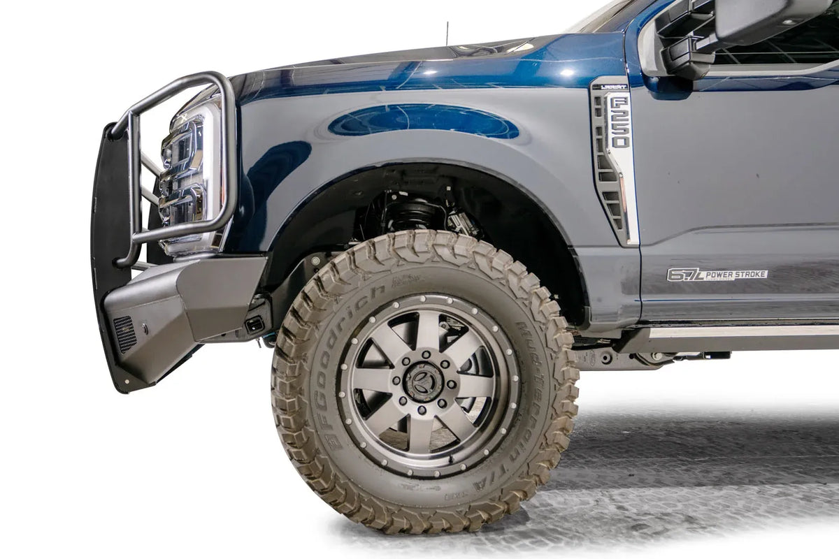 Fab Fours Black Steel Elite front bumper with full guard on a dark blue Ford Super Duty, driver-side profile view of the front corner, showing full grille guard uprights and side hoops, the smooth steel face, mesh-backed side openings, and the center sensor/plate mount