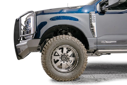 Fab Fours Black Steel Elite front bumper with full guard on a dark blue Ford Super Duty, driver-side profile view of the front corner, showing full grille guard uprights and side hoops, the smooth steel face, mesh-backed side openings, and the center sensor/plate mount