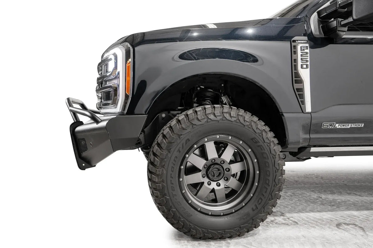 Fab Fours Black Steel Elite front bumper with pre-runner guard on a dark blue Ford Super Duty, driver-side profile view of the front corner, showing the tubular pre-runner hoop, the smooth steel face, mesh-backed side openings, and the center sensor/plate mount