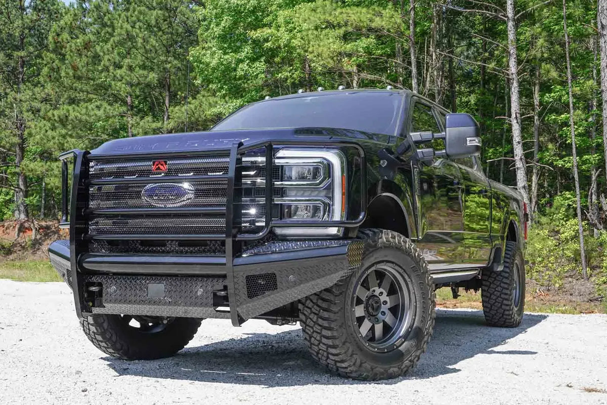 Fab Fours Black Steel front bumper with full guard on a black Ford Super Duty, front driver-side three-quarter view outdoors, showing full grille guard with mesh insert, diamond-plate face panels, the center sensor/plate mount, and angled side wings