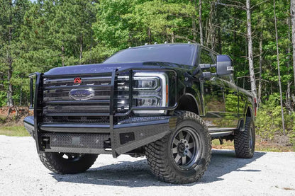 Fab Fours Black Steel front bumper with full guard on a black Ford Super Duty, front driver-side three-quarter view outdoors, showing full grille guard with mesh insert, diamond-plate face panels, the center sensor/plate mount, and angled side wings
