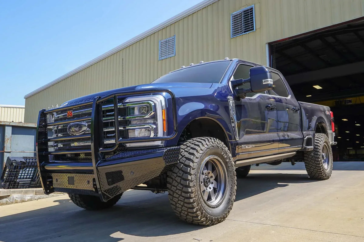 Fab Fours Black Steel front bumper with full guard on a blue Ford Super Duty, front driver-side three-quarter view outdoors, showing full grille guard with mesh insert, diamond-plate face panels, the center sensor/plate mount, and angled side wings