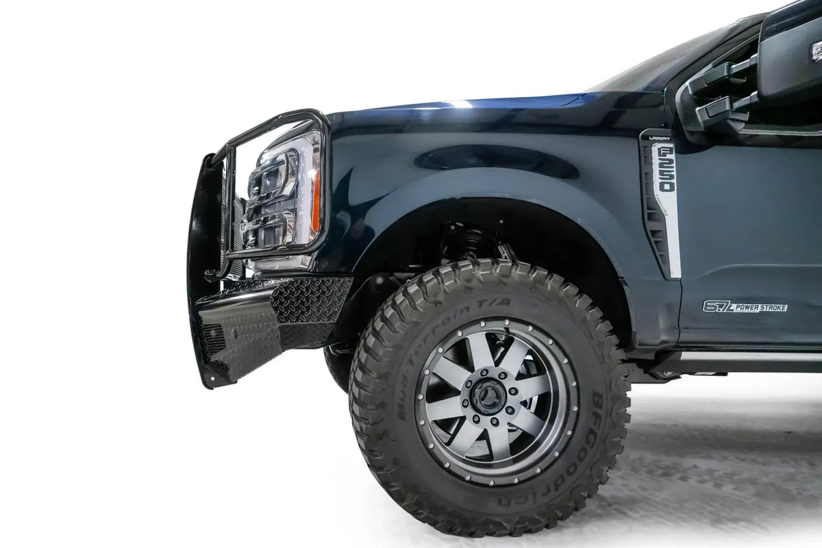Fab Fours Black Steel front bumper with full guard on a dark blue Ford Super Duty, driver-side profile view of the front corner, showing full grille guard with mesh insert, diamond-plate face panels, the center sensor/plate mount, and angled side wings
